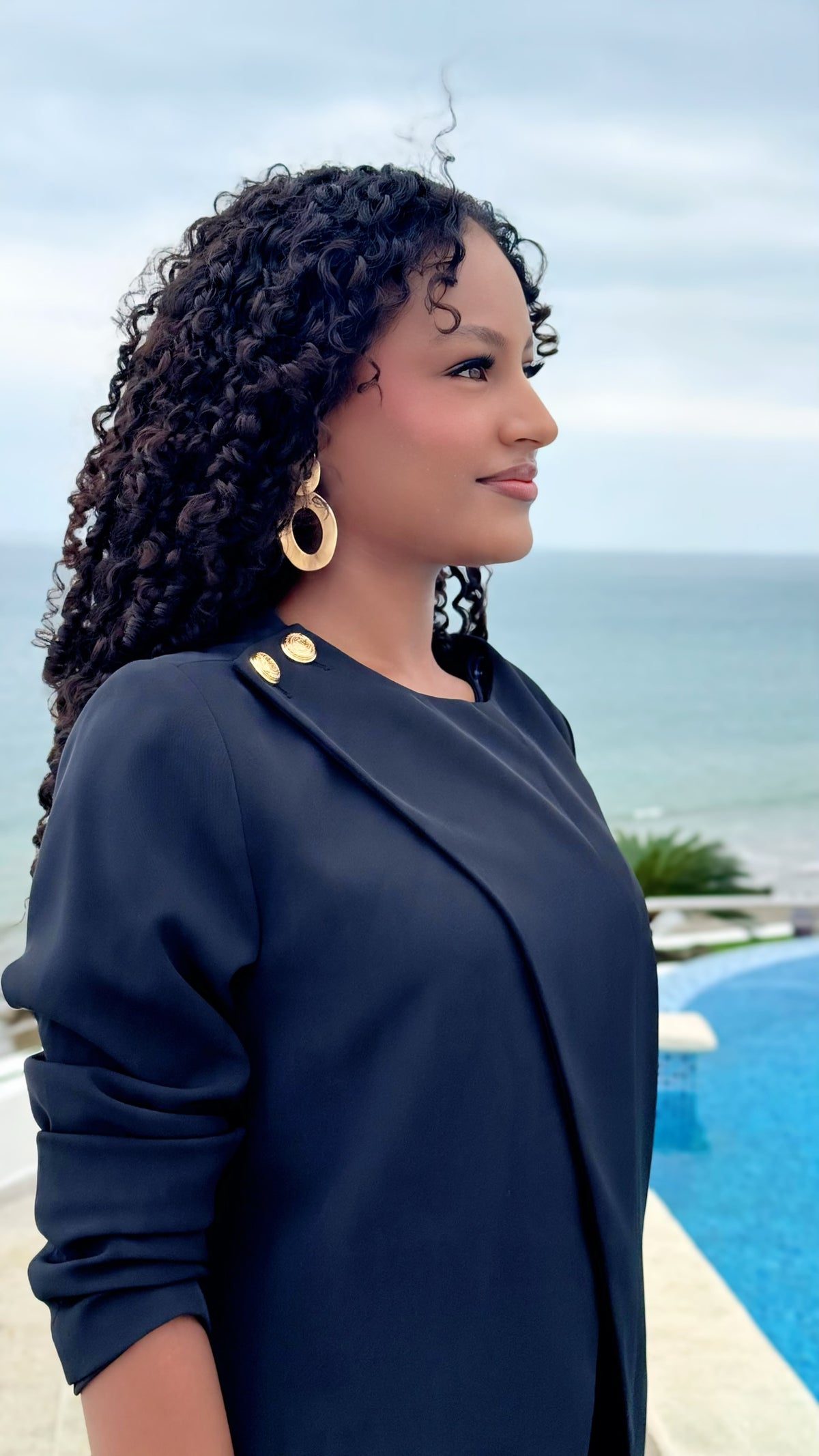 Mujer con blazer negro Exotica de corte cruzado y botones dorados, estilo formal frente al mar.
