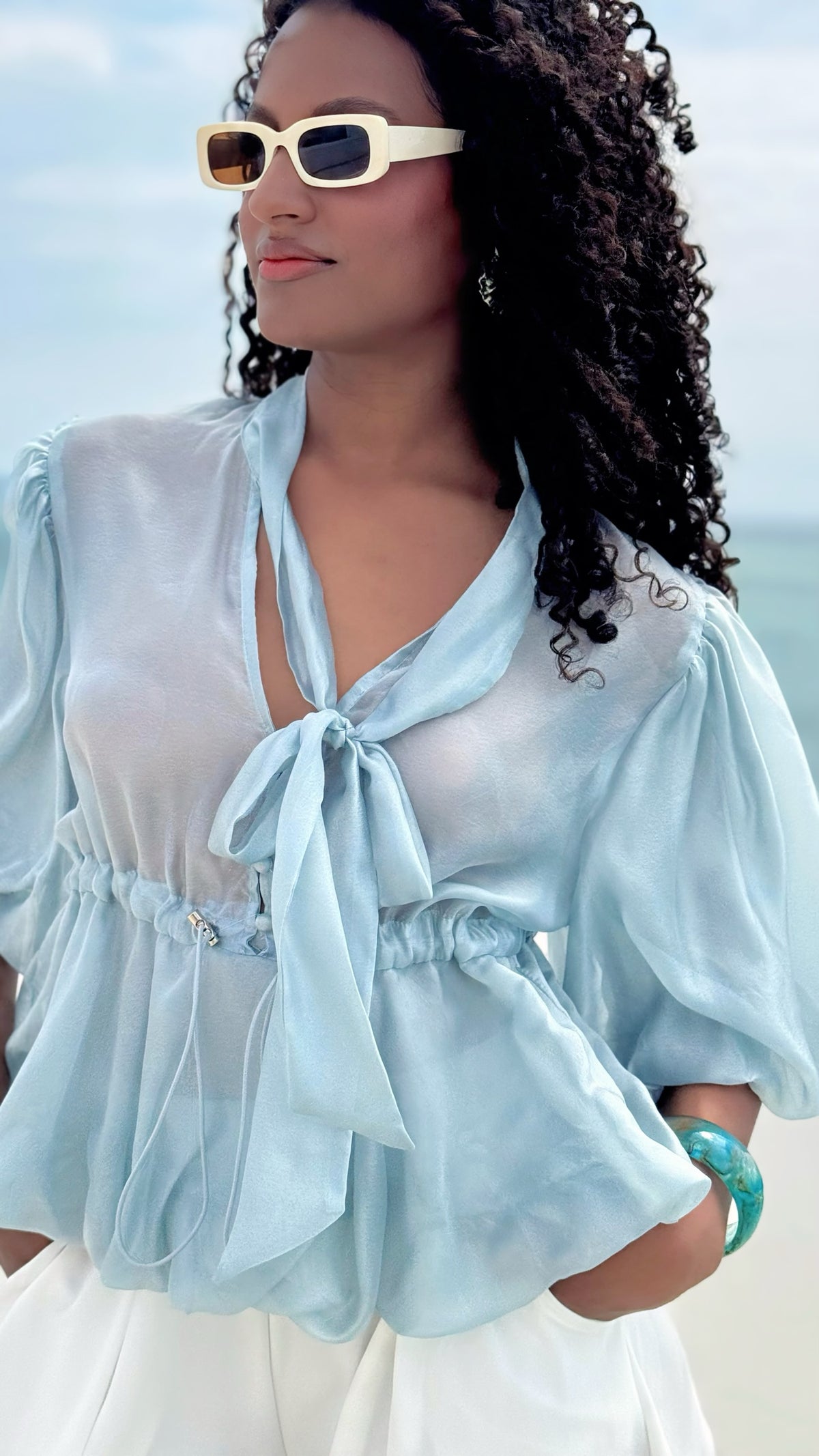 Modelo con blusa Exotica de organza azul cielo, mangas abullonadas, lazo al cuello y cintura ajustable en la playa.