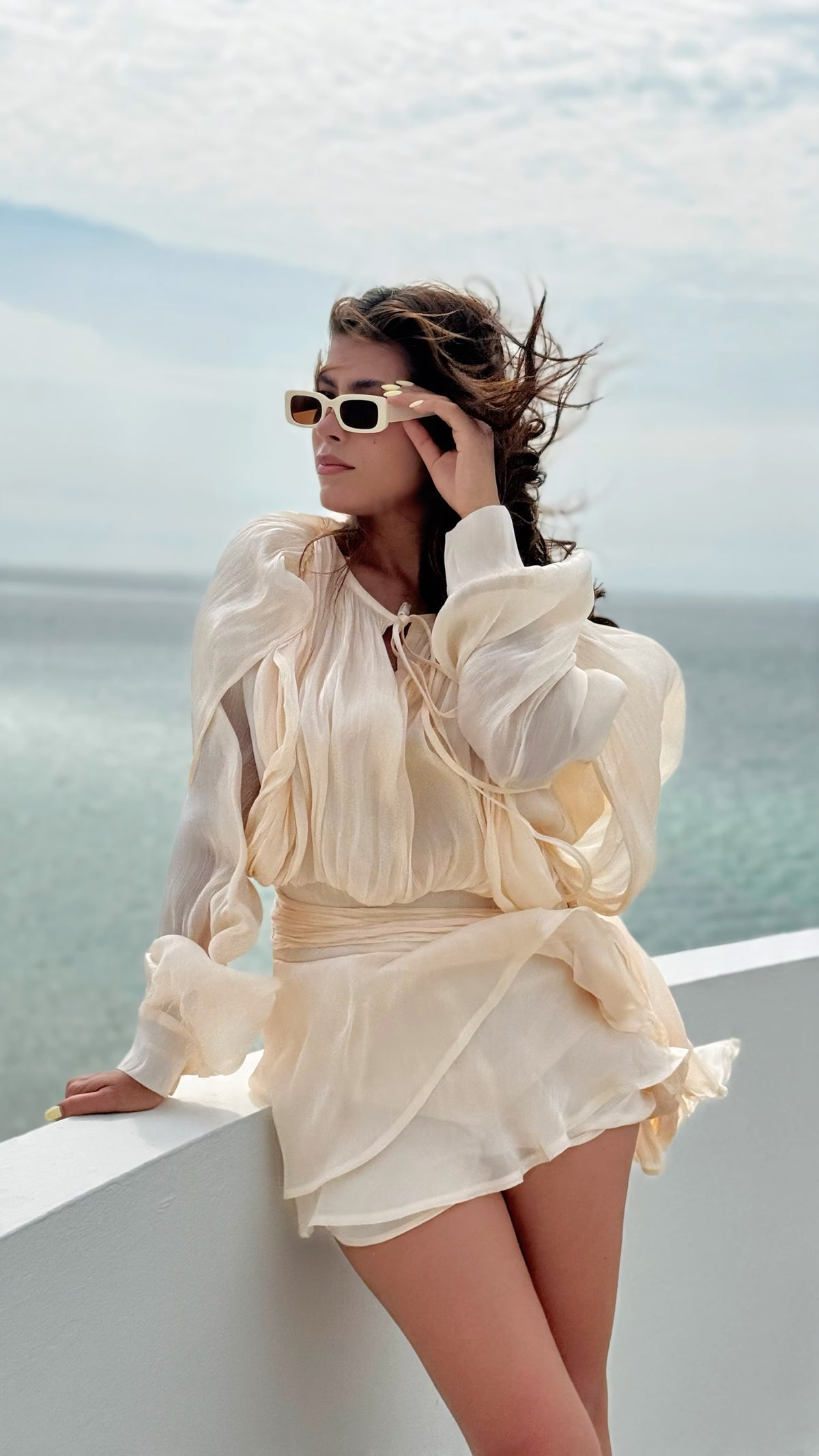 Exotica champagne sheer mini dress with bishop sleeves and tie-neck detail worn by a woman at the coast.