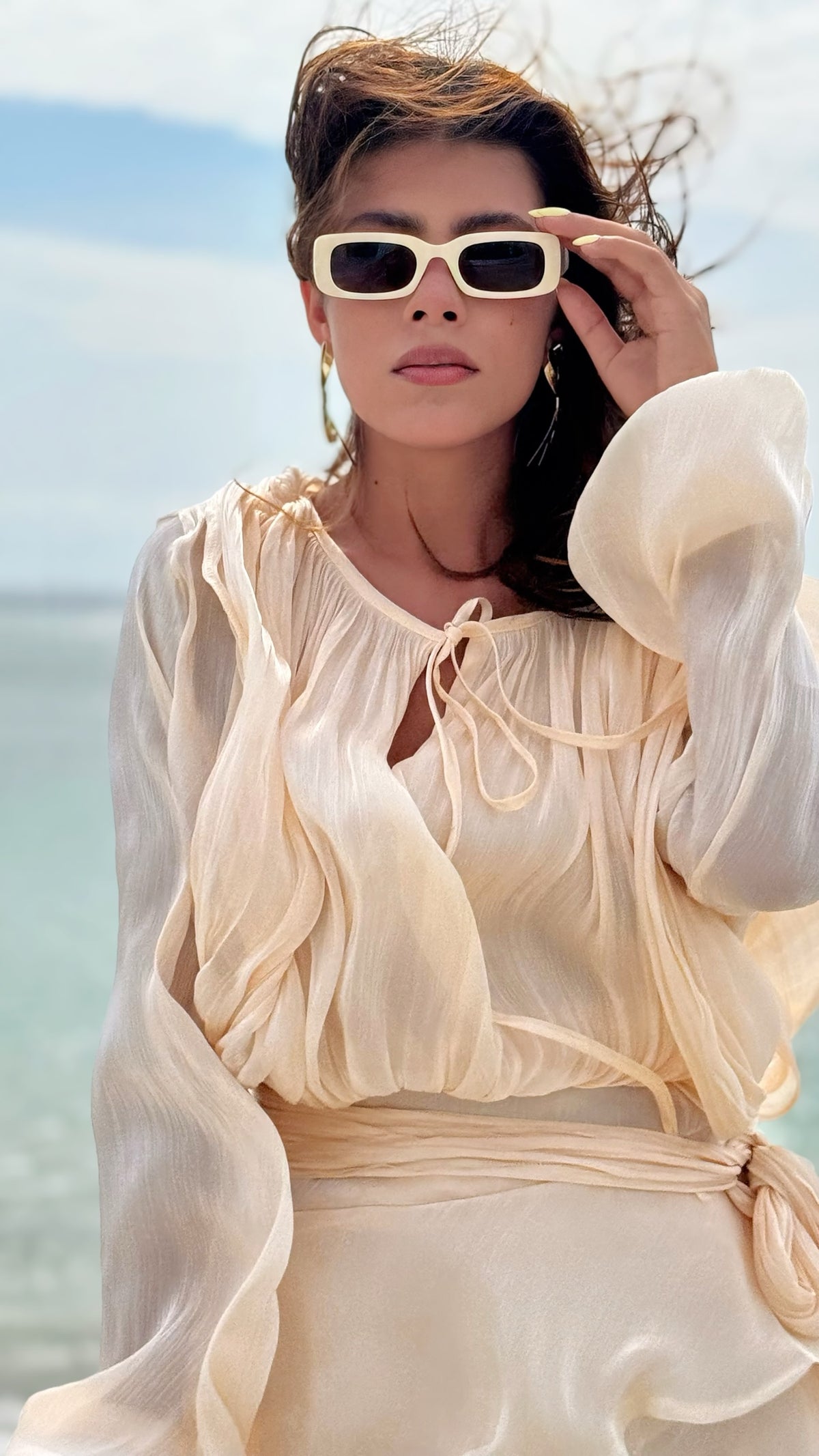 Exotica champagne sheer mini dress with bishop sleeves and tie-neck detail worn by a woman in sunglasses at the beach