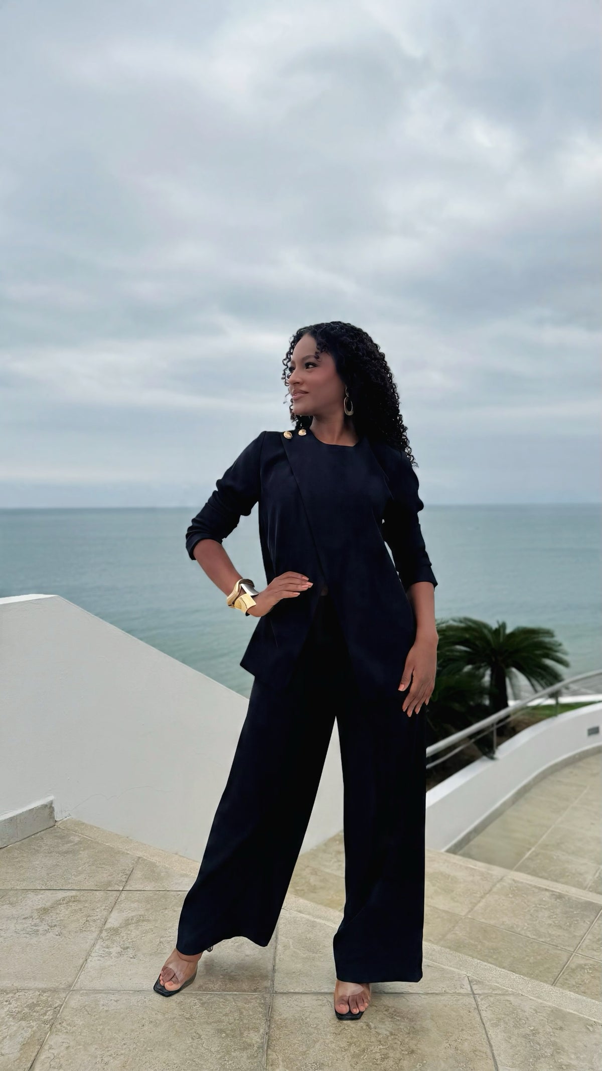 Mujer con conjunto formal Exotica negro de blazer cruzado con botones dorados y pantalón recto frente al mar.