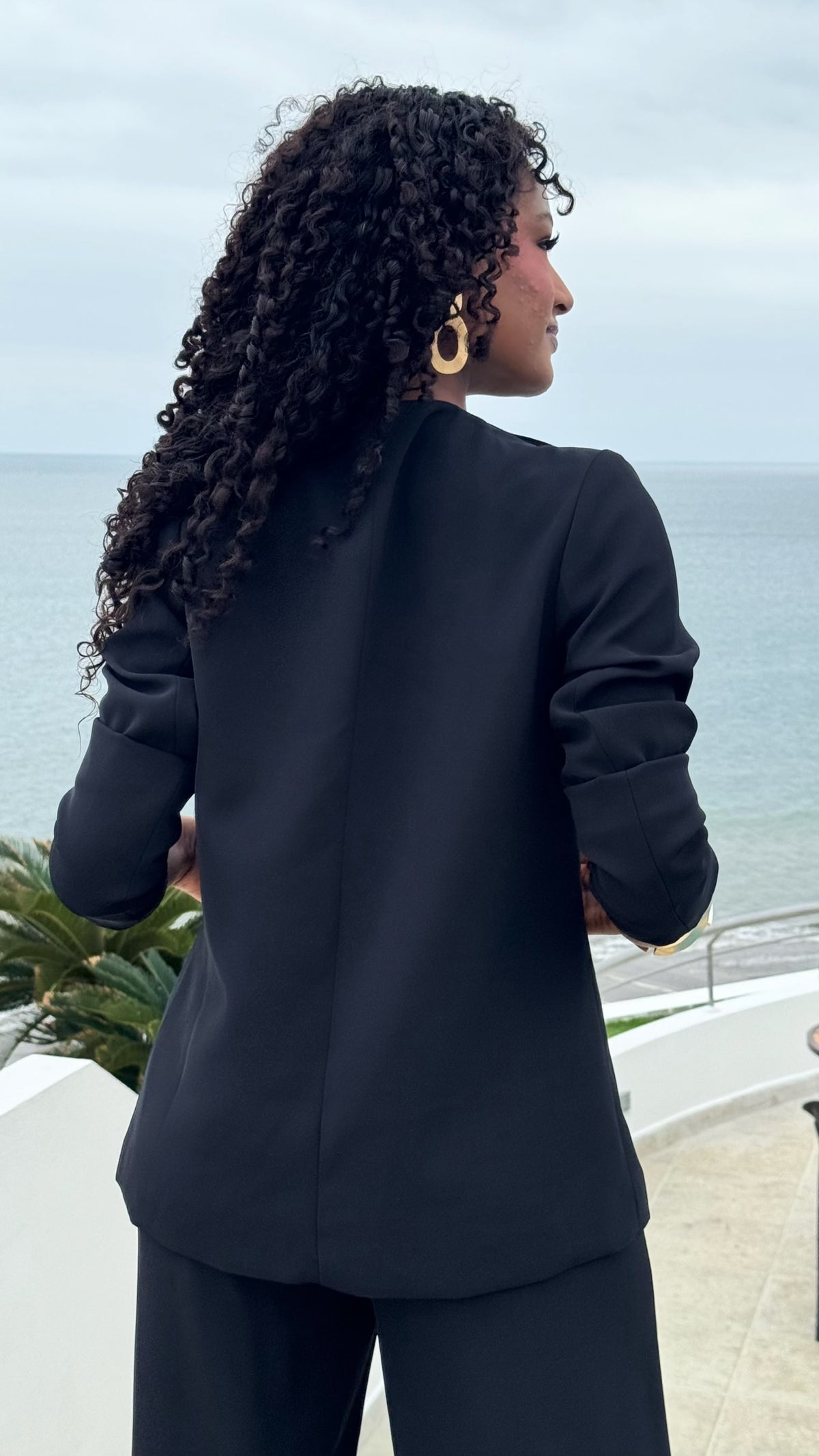 Mujer con blazer negro Exotica de corte estructurado y pantalón a juego, vista posterior frente al mar
