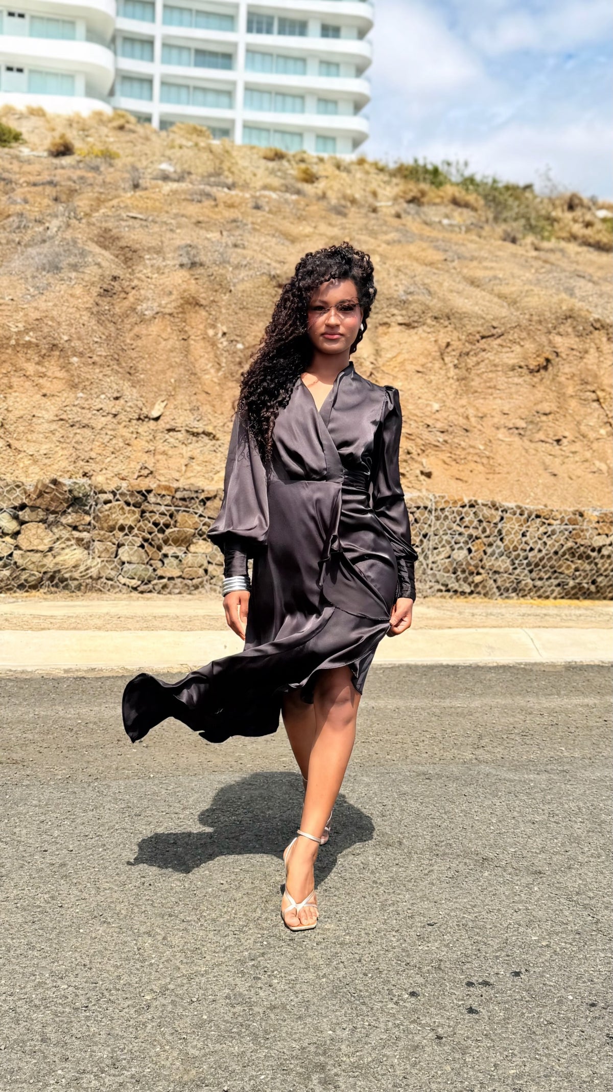 Modelo con vestido midi cruzado de satén negro Exotica, mangas largas abullonadas y bajo con volantes en exterior.